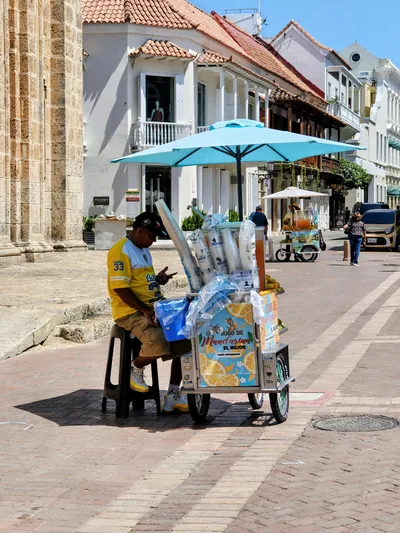 An ice cream vendor sits on a street in Cartagena, Colombia, under a sunny sky.