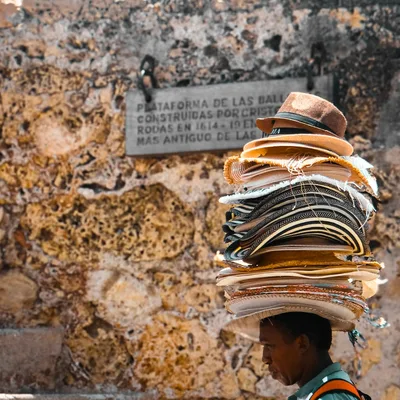 A street vendor in Cartagena balances a stack of hats against a historic stone wall.
